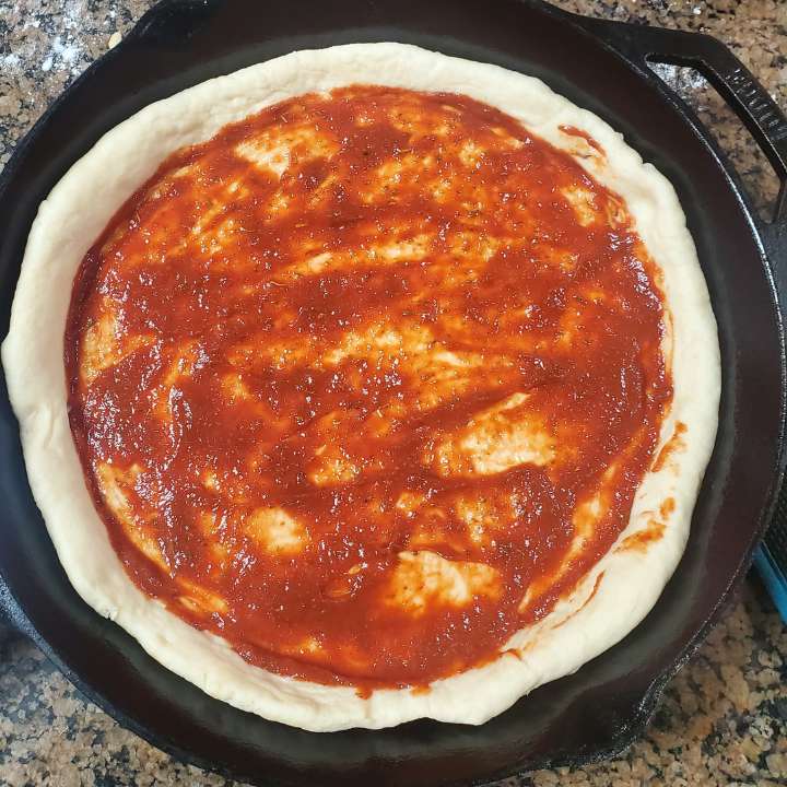 Black cast iron frying pan with pizza dough inside with twinethyme homemade pizza sauce spread out on the crust.